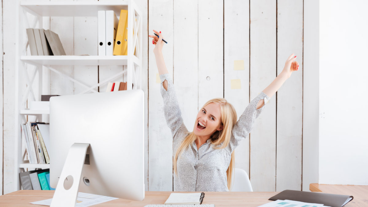 Happy excited business woman celebrating success while sitting at her workplace with hands raised